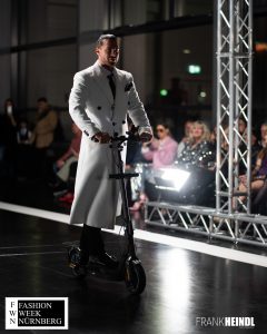 Markus in weißem zwei Reihen-Mantel (britischer Stil) mit E-Roller auf dem Laufsteg der Fashionweek Nürnberg seitliche Ansicht - LikeMan Maßanzüge Nürnberg