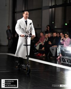 Markus in weißem zwei Reihen-Mantel (britischer Stil) mit E-Roller auf dem Laufsteg der Fashionweek Nürnberg von vorne - LikeMan Maßanzüge Nürnberg