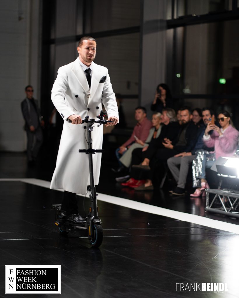 Markus in weißem zwei Reihen-Mantel (britischer Stil) mit E-Roller auf dem Laufsteg der Fashionweek Nürnberg von vorne - LikeMan Maßanzüge Nürnberg