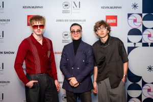 Morteza Faizi Designer blickt in Kamera auf der X-Mas Fashionweek München vor der Wand mit 2 Models - LikeMan Maßanzüge Nürnberg