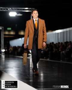 Reiner in braunem Mantel mit dunkelgrauem Anzug (britischer Stil) auf dem Laufsteg der Fashionweek Nürnberg seitliche Ansicht - LikeMan Maßanzüge Nürnberg